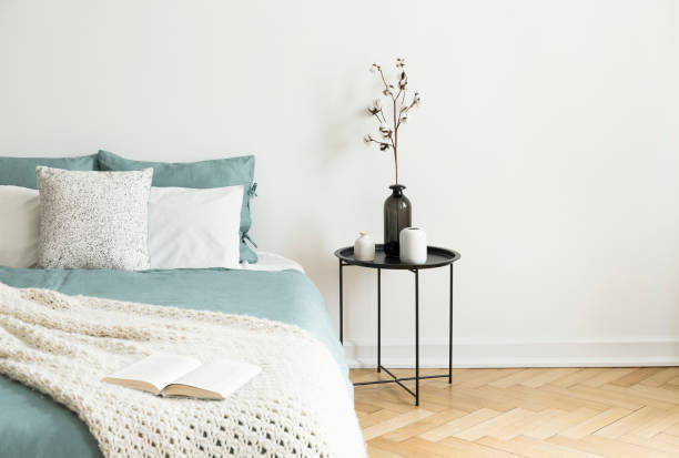 istock Black table next to bed with green sheets, pillows and knit blanket in white bedroom interior. Real photo 1007742586
