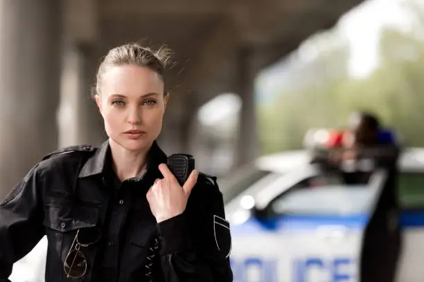 policewoman using walkie-talkie and looking at camera with blurred partner near car on background