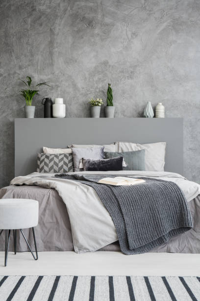istock Front view of a comfortable bed with linen, pillows, a blanket and an open book on it in a grey style bedroom interior. Green plants and vases as decorations. Real photo 1007782218