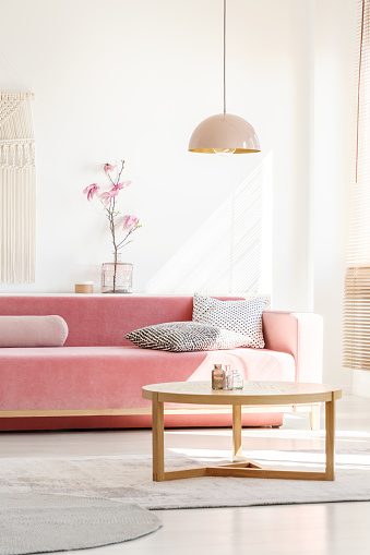 Estilo retro, lámpara colgante rosa milenaria de encima de una mesa sencilla de madera en un interior de salón soleado, blanco con estampado almohadas en un sofá de terciopelo photo