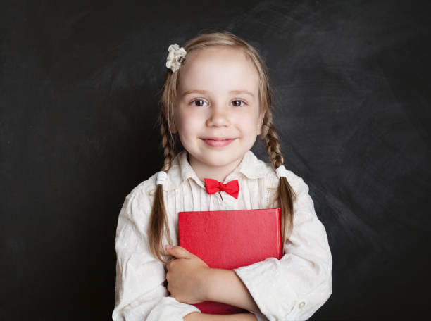 istock Cute child girl with book having fun on chalkboard background in elementary school classroom. Back to school, reading and education concept 1007786732