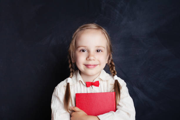 istock Smiling girl with red book on empty chalkboard background with copy space. Child portrait 1007787218