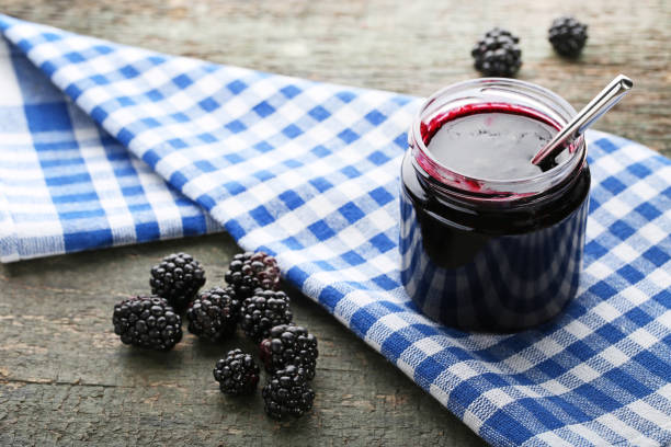 Blackberry jam in glass jar with spoon and napkin on grey wooden table stock photo