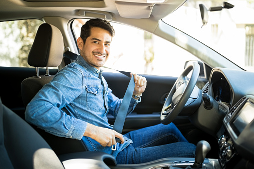 Portrait of young hispanic man sitting in driving seat of car, fastening safety belt and making en eye contact