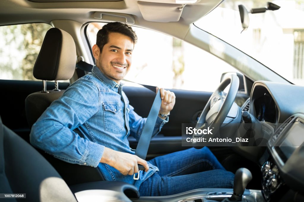 Man sitting in car seat fastening seat belt Portrait of young hispanic man sitting in driving seat of car, fastening safety belt and making en eye contact Driving Stock Photo