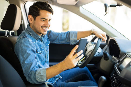 Portrait of young male driver using his mobile phone while driving the car and smiling