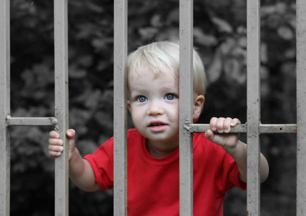 Cute upset blond toddler boy behind bars. Parenting difficulties or juvenile justice concept.