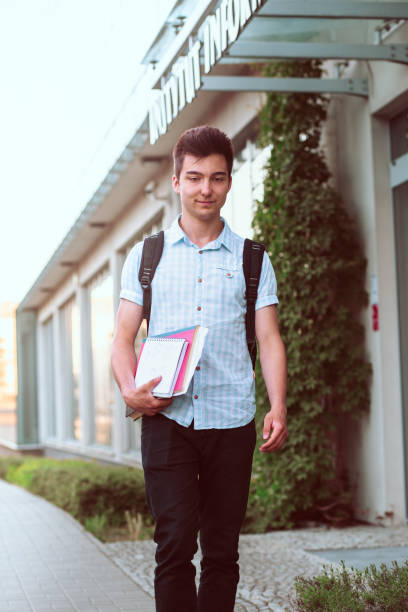 istock Student holding a notebook and carrying a backpack walking at the front of university building. Young smiling boy wearing blue shirt and dark jeans 1007964384