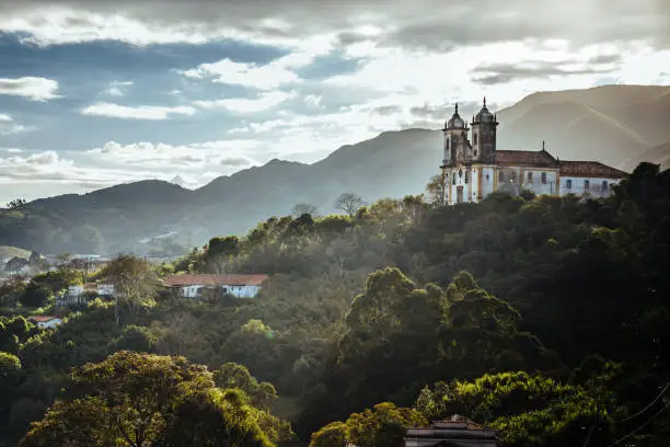 Landscape of Ouro Preto city of Minas Gerais, Brazil with Sao Francisco de Paula Church in the top of a mountain under sunshine and dark clouds full of green trees and fog.