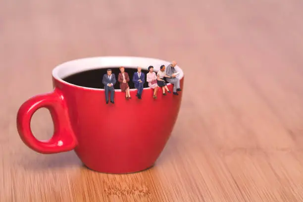 Creative concept about drinking coffee and waiting. Miniature people sit on the edge of a cup of coffee tea coffee break. Red cup on a wooden background.