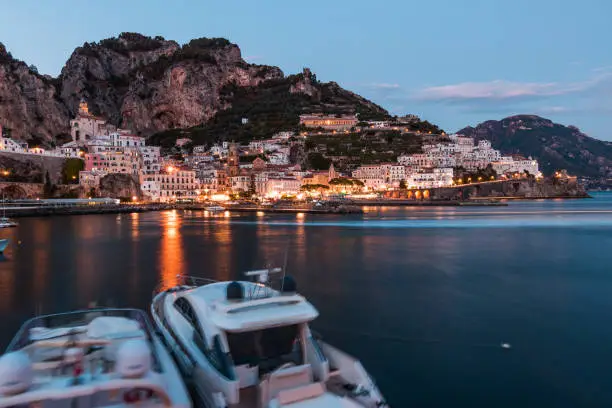 Long exposure shot taken at sunset in Amalfi, Italy. Water reflections of city lights can be clearly seen. Mountains of the amalfi coast and the town can also be seen in the background.