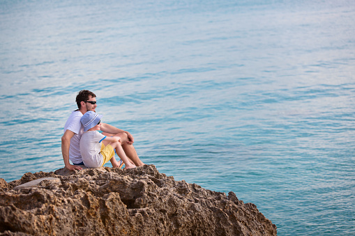 family of two, father and son, enjoying sunset together sitting at the rock at anguilla island, caribbean, copy space on right