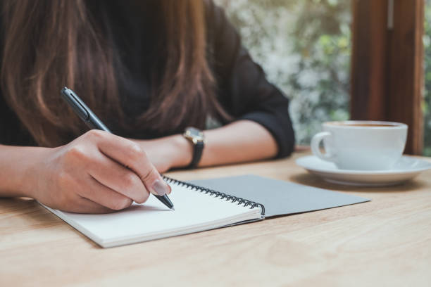 istock Closeup image of a woman's hand writing down on a white blank notebook with coffee cup on wooden table 1008161388