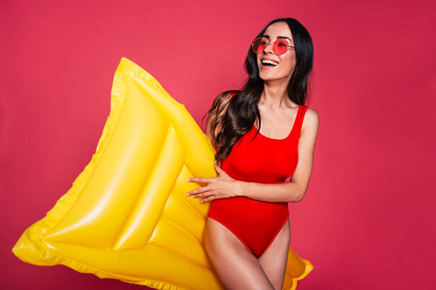 bella giovane donna sorridente vestita con costume da bagno rosso e occhiali in posa con materasso gonfiabile giallo isolato su sfondo rosa - swimwear bikini women fashion model foto e immagini stock