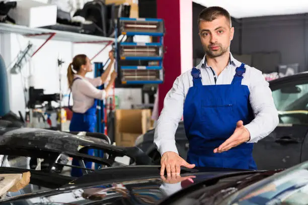 Serious male worker in blue overalls showing car after repainting at auto service