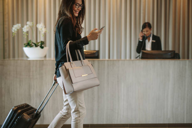 Business traveler in hotel hallway with phone Woman using mobile phone and pulling her suitcase in a hotel lobby. Female business traveler walking in hotel hallway. reception desk photos stock pictures, royalty-free photos & images