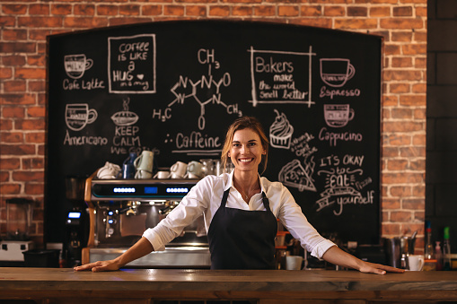 Portrait of confident female barista standing behind counter. Woman cafe owner in apron looking at camera and smiling.