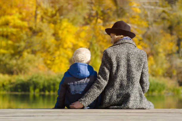 istock Blond boy and his mother sitting on the dock. Autumn day. Back view 1008311770