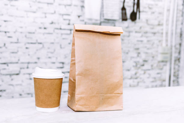 Cup of coffee and blank paper bag with copy space for your brand on table against white brick wall background stock photo