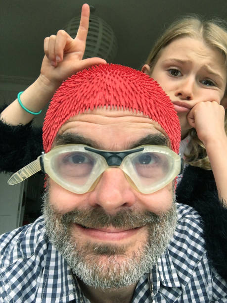 Loser Dad selfie - unimpressed photobomber daugther makes an L sign at father Young girl photobombing making loser L sign with her hand as her father takes selfie in swimming goggles and rubber swimming cap photo bomb stock pictures, royalty-free photos & images