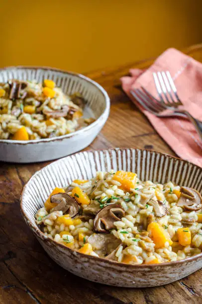 autumn risotto with butternut squash and porcini mushrooms garnished with rosemary in rustic homemade pottery with orange napkin and forks on a warm wooden table