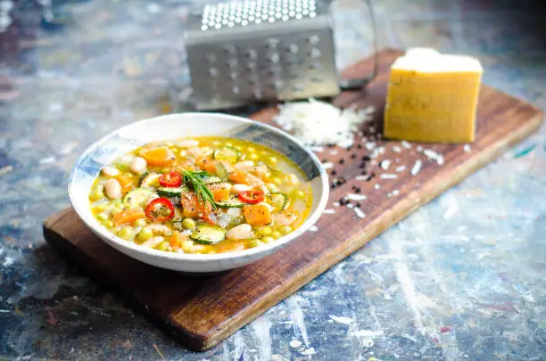 earthy minestrone soup with fresh vegetables in broth. zucchini, carrots, peas, cannellini beans, onion, spring onion, red chilli garnished with rosemary in a stylish rustic bowl on a wooden serving board with parmesan cheese and grater and pepper grains in the background