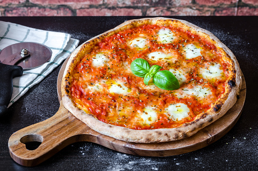 classic pizza margherita made with a sourdough base, tomato sauce with oregano and olive oil, mozzarella cheese and fresh basil on a wooden board, black table, cutting wheel, towel and brick wall background