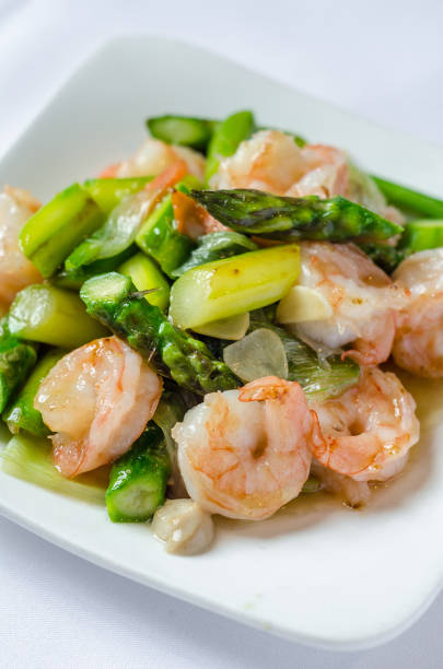 Close-up composition showcasing parallel lines of grilled jumbo shrimp, steamed asparagus spears, and fresh lemon slices artfully arranged on textured gray slate board.