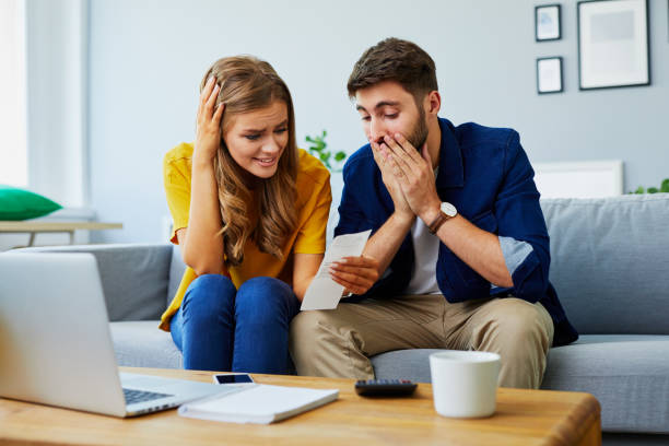 vista frontal de una joven pareja estresada frente a problemas financieros, sentado en el sofá y mirando facturas - crisis european union currency europe debt fotografías e imágenes de stock