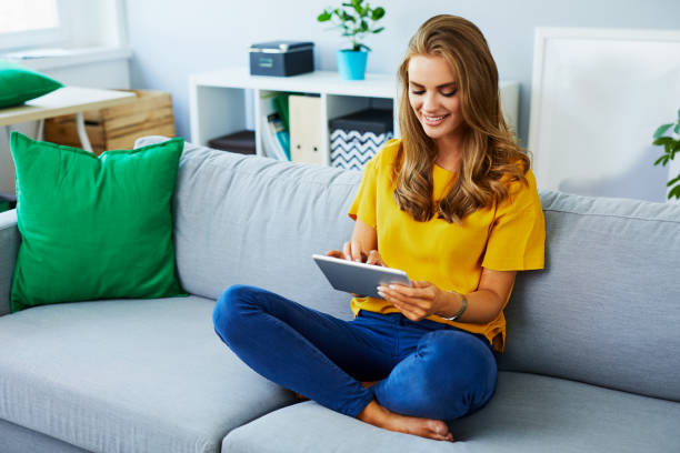 istock Beautiful young woman sitting on the sofa at home and using tablet, touching the screen and smiling 1008375572