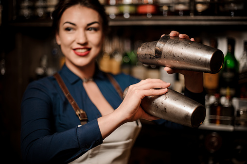 Attractive bartender girl in the white apron holding in her hands two steel cocktail shakers at the bar counter