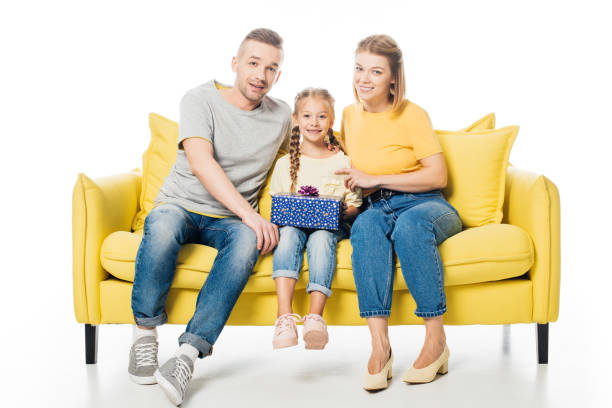 istock smiling parents and daughter with wrapped gift sitting on yellow sofa isolated on white 1008415426