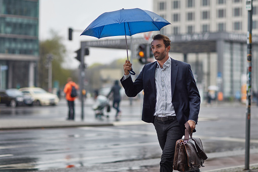 Mid adult businessman holding umbrella while walking on sidewalk. Male executive is carrying bag in rain. He is in formals at city.