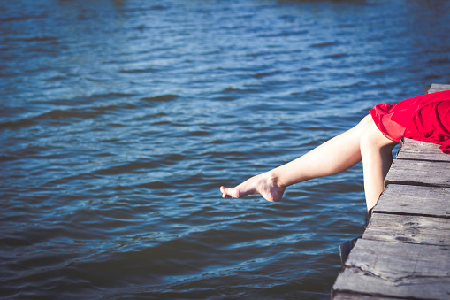 Legs of a young Caucasian woman wearing red dress while she's sitting on a wooden jetty on a lake on a sunny summer day.