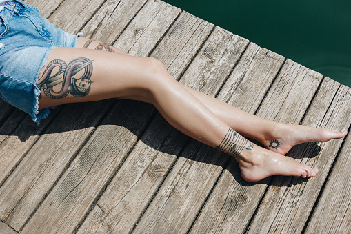 cropped shot of barefoot girl in denim shorts resting on wooden pier near water