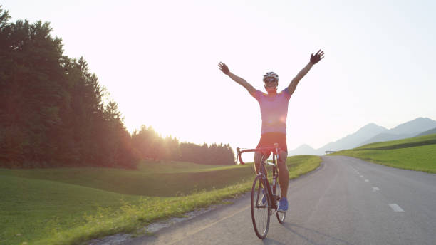 LENS FLARE: Happy sportsman riding his bicycle victoriously outstretching arms. LENS FLARE: Happy sportsman riding his bicycle victoriously outstretching his arms while cruising through tranquil sunlit nature on gorgeous summer afternoon. Thrilled bicycle rider celebrates win. super bike stock pictures, royalty-free photos & images