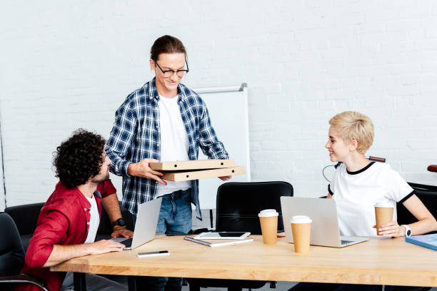 istock smiling young man holding pizza boxes while colleagues sitting at table in office 1008581452