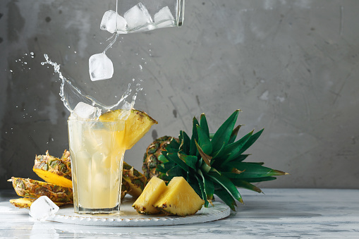 Pineapple fresh juice with falling ice cubes making splash. Cut fruit slices on wooden table and grey concrete background, front view