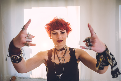 Young red haired woman dressed in rock and heavy metal style raising her hands and showing devil horns gesture