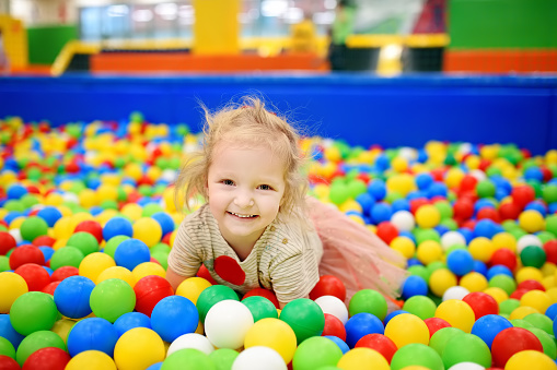 Curly little girl having fun in ball pit with colorful balls. Child playing on indoor playground. Kid jumping in ball pool.