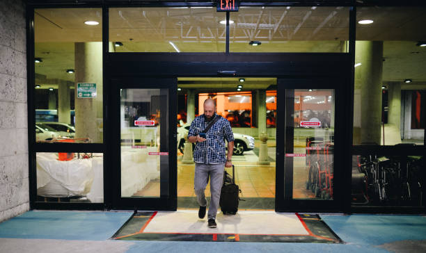 Airport hustle Man walks through the sliding glass doors of an airport, he pulls a suitcase and wears a carry on strap. He is dressed casually and has a beard, is looking at his phone, perhaps working from his phone. walking into glass door stock pictures, royalty-free photos & images
