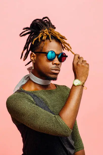 Portrait of a young man in green with dreadlocks and blue sunglasses, isolated on pink