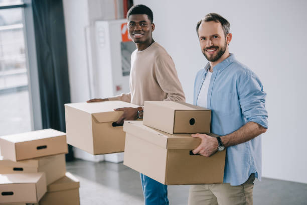 multiethnic coworkers holding cardboard boxes and smiling at camera during relocation stock photo