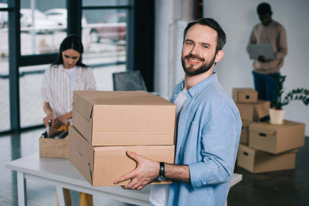istock man holding cardboard boxes and smiling at camera while relocating with colleagues at new workplace 1008845052