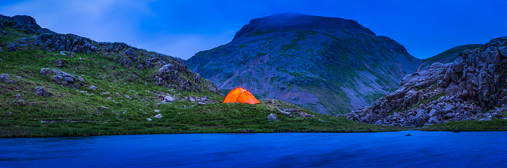 Brightly coloured dome tent warmly illuminated against the blue dusk landscape and lakeside of a wilderness camp high in the mountains of the Lake District, Cumbria, UK.