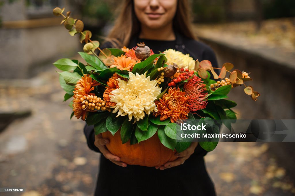 Woman holding a pumpkin with autumn flowers Smiling woman in black dress holding a beautiful pumpkin with beautiful autumn flowers on the outdoors background Autumn Stock Photo