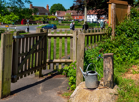 Some people walking on the village green past the graveyard of St Bartholomew's Church in the pretty village of Otford in Kent, Southeast England, on a sunny spring day, where an old-fashioned metal watering can stands on a drain cover beneath a water tap.