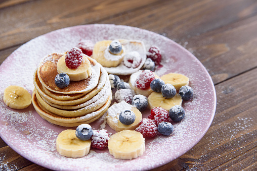 Homemade delicious pancakes with fresh berries sprinkled with powdered sugar in a violet plate on wooden background. A tasty and healthy breakfast of pancakes with raspberries and blueberries.