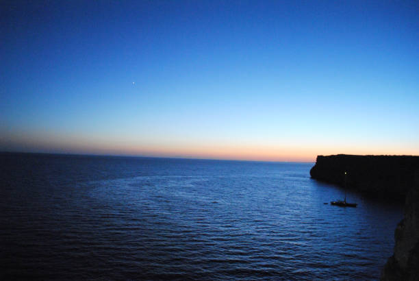 Beautiful sunset at Cova d'en Xoroi caves on island of Menorca. Seashore with cliffs and sailing boat parked in bay.