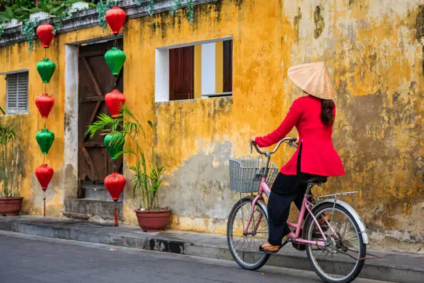 Vietnamese woman riding a bicycle in old town in Hoi An city, Vietnam. Hoi An is situated on the east coast of Vietnam. Its old town is a UNESCO World Heritage Site because of its historical buildings.